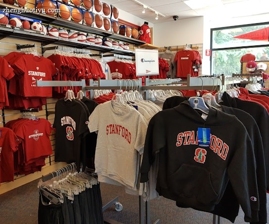 A retail display in a store features Stanford University merchandise, including red and grey t-shirts, hoodies, and sports equipment like basketballs and footballs on shelves and racks.
